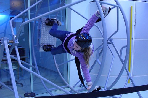 Ein Mädchen in Jeans und einem violetten Pullover ist in ein Gerät ähnlich einer Zentrifuge eingebunden. Sie trägt einen Helm. Sie befindet sich in einer waagerechten Schwebeposition.