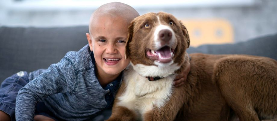 Ein Kind sitzt gemeinsam mit einem Hund auf dem Sofa und lächelt gutgelaunt in die Kamera.