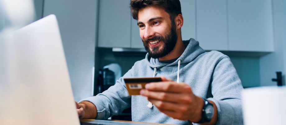 Mann sitzt mit Laptop am Küchentisch und hält Bankkarte in der Hand