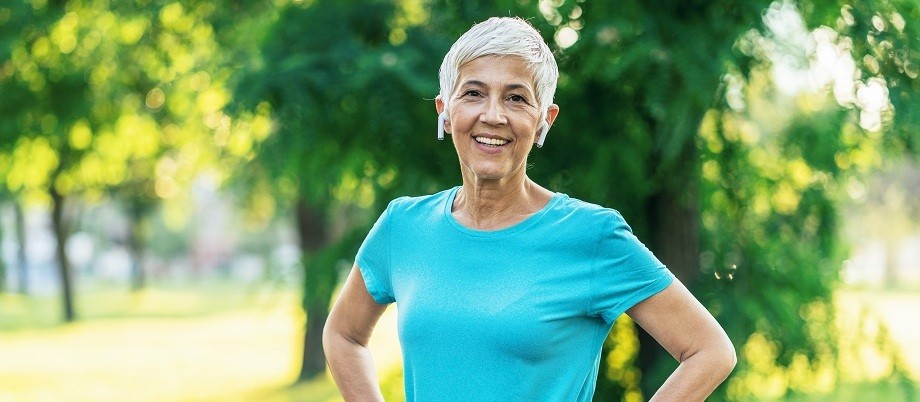 Gesundheitsvorsorge ab 50: Eine sportliche Frau reiferen Alters im Park macht eine kurze Pause vom Joggen und lächelt, die Hände in die Hüfte gestemmt, in die Kamera.