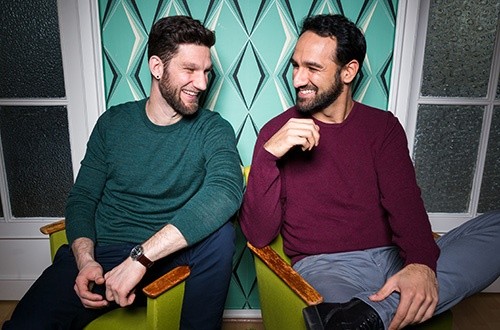 Two young men, smiling, in front of a patterned wallpaper. 