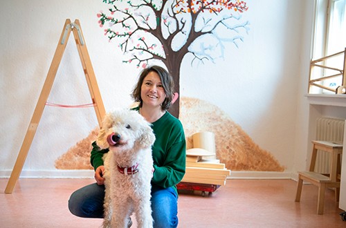 Ein Hund mit weißem Fell steht in der Bildmitte. Dahinter sitzt eine Frau in Jeans und grünem Pullover. An der Wand im Hintergrund ist ein Baum aufgemalt.
